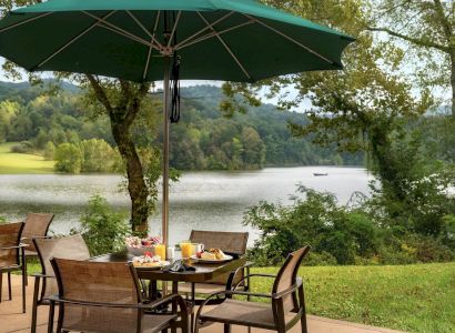 An outdoor dining area with a table under a green umbrella, set with meals, overlooking a serene lake surrounded by trees and hills in the background.