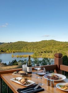 A table with wine and food on a balcony overlooks a scenic view of a lake and lush green hills under a clear blue sky.