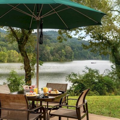 An outdoor patio with a table set for a meal under a green umbrella, surrounded by chairs, overlooking a serene lake with lush greenery.