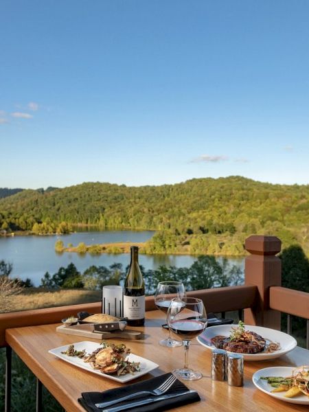 A table with a meal set for two overlooking a scenic lake and tree-covered hills, featuring wine, main courses, and side dishes.