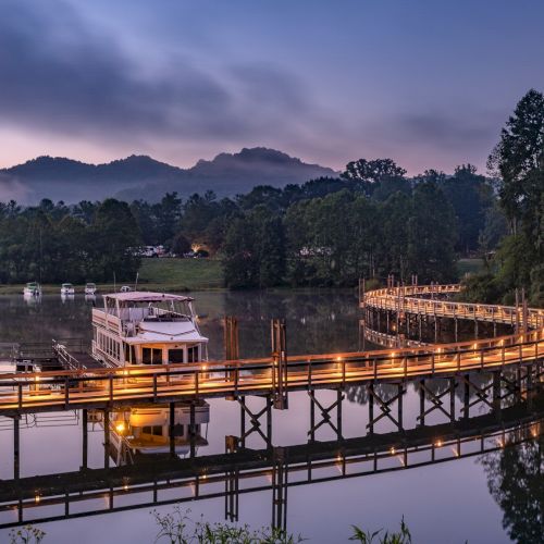 A wooden pier with lights curves over a calm lake, surrounded by lush greenery and mountains in the background under a twilight sky.