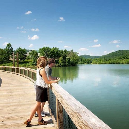 A couple stands on a wooden walkway, overlooking a serene lake surrounded by green hills under a clear blue sky with scattered clouds.