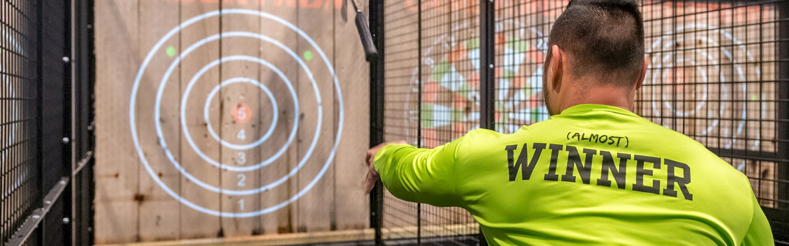 A person in a bright green shirt is throwing an axe at a target in an enclosed area. The shirt has the word 