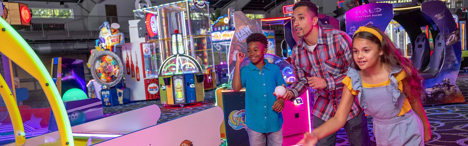 A family of four plays a colorful arcade game at an indoor amusement center, with glowing lights and arcade machines in the background.