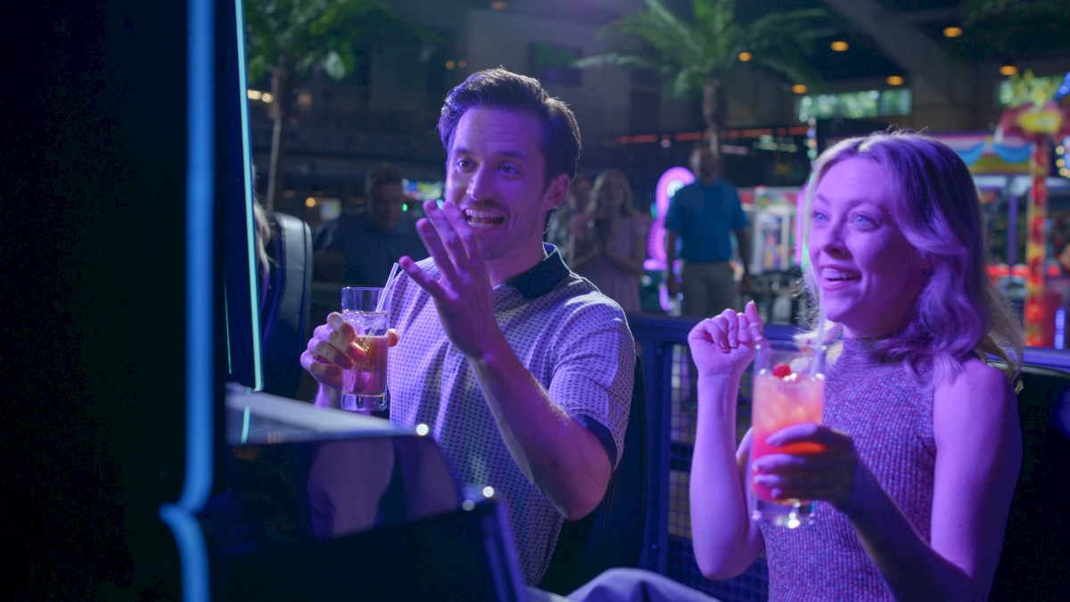 Two friends play arcade games at a neon-lit arcade bar, holding drinks and smiling as they compete.
