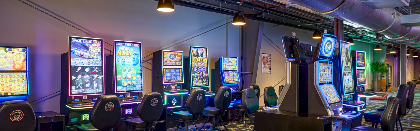A row of arcade gambling machines with stools along a neon-lit gaming room, large vents overhead and circular patterned carpet.