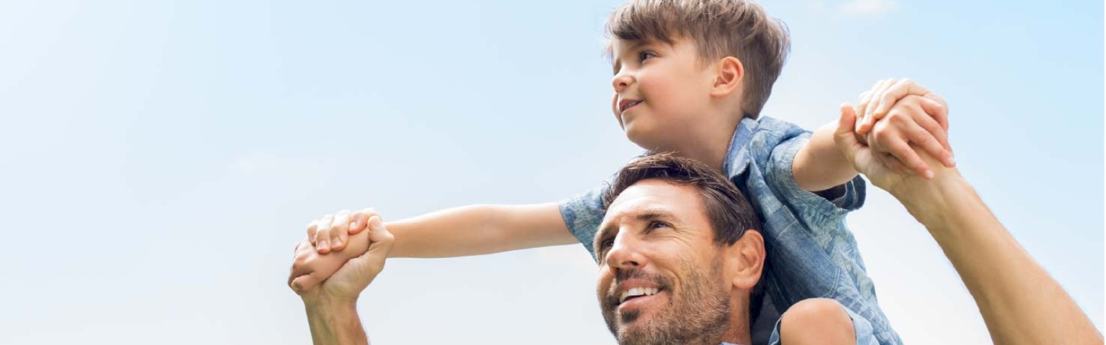 A man is carrying a child on his shoulders against a blue sky, holding hands and smiling.