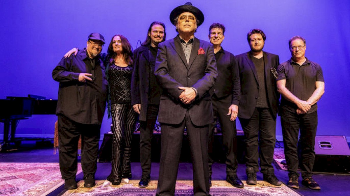 A group of seven people dressed in dark clothing stands on a stage, with a man in front wearing a hat and suit.