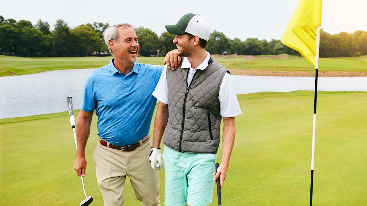 Two people walking on a golf course, holding clubs, and smiling at each other near a flagstick.