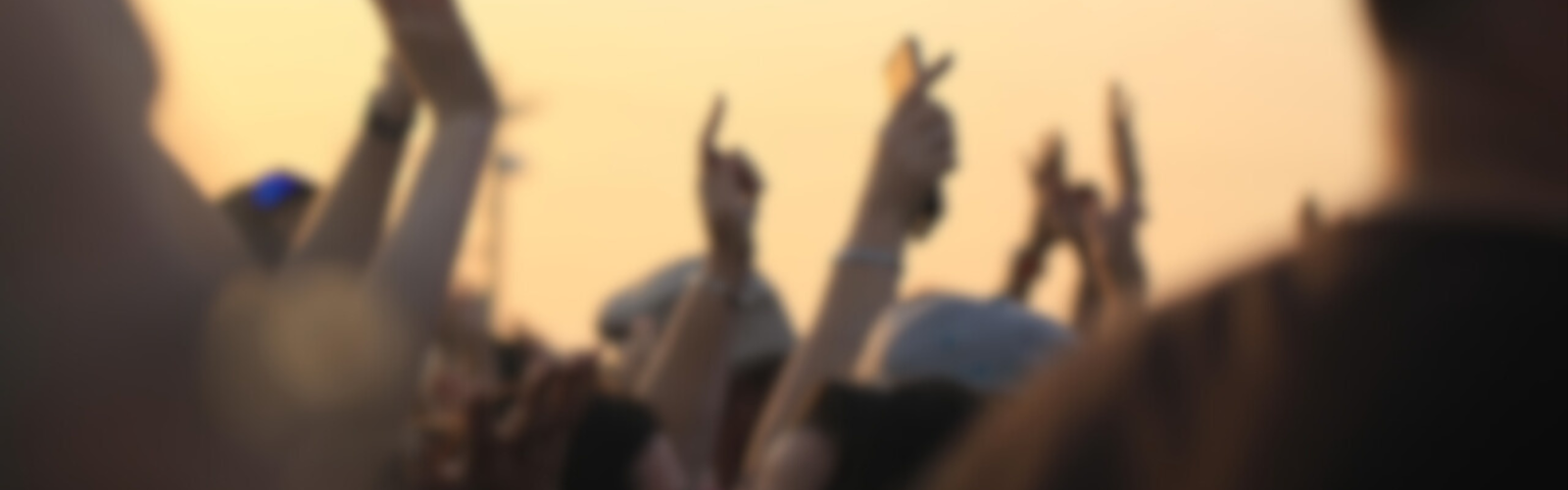 Blurry image of a crowd outdoors, hands raised, likely at a concert or event during sunset.