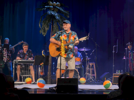 A man in a Hawaiian shirt and gray shorts plays guitar on stage decorated with beach balls, surrounded by band members, under colorful lighting.