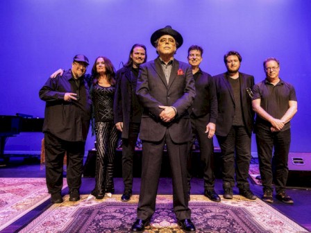 A group of seven people in dark attire on stage, one person in the center wearing a hat, standing on a patterned rug.