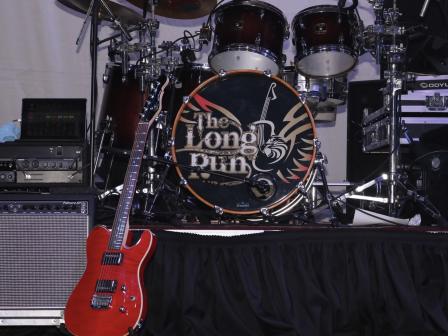 A red electric guitar rests against an amplifier, in front of a drum set with 