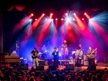 A band performs on a stage with colorful lights overhead and a crowd watching in a concert setting.
