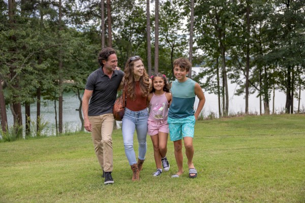 A family of four is happily walking on a grassy field surrounded by trees and a lake.