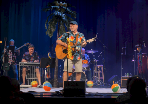 A group of musicians performing on stage with tropical decorations and beach balls, creating a lively, summer-themed atmosphere.