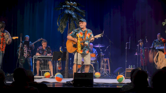 A group of musicians performing on stage with tropical decorations and beach balls, creating a lively, summer-themed atmosphere.