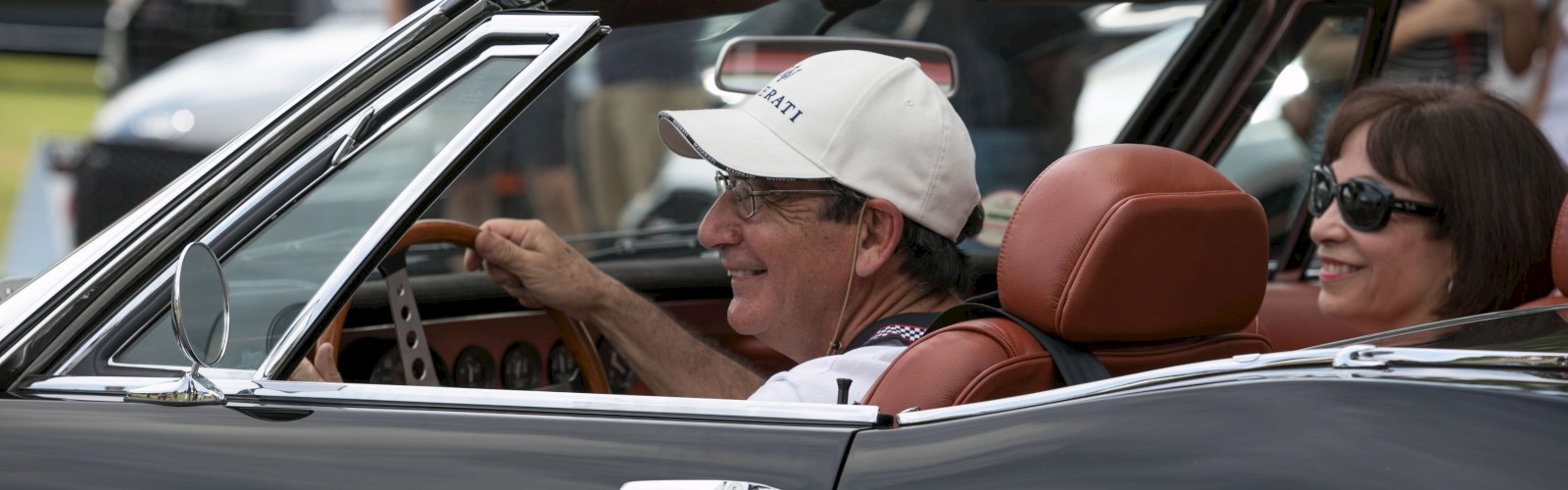 A man and woman are smiling while driving a classic black convertible car in a lively outdoor setting with people in the background.