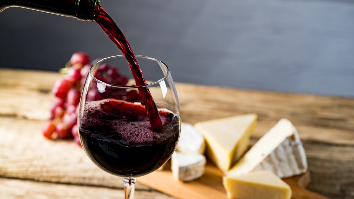 A glass of red wine is being poured on a wooden table with a platter of assorted cheeses and grapes in the background.
