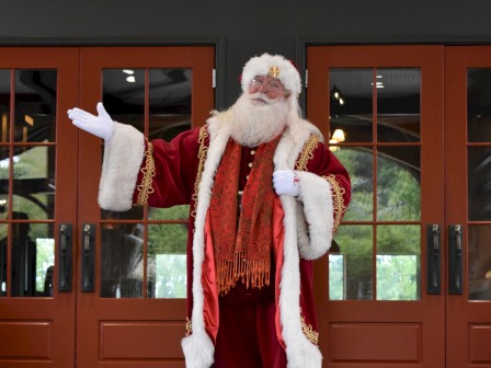 A person in a Santa Claus outfit stands in front of wooden doors, with arms open wide, on a tiled surface.