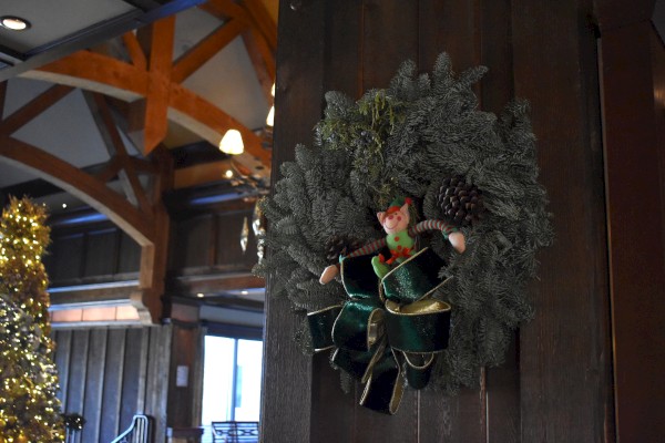 A festive wreath with a green bow, pine cones, and a small elf doll hanging on a wooden wall indoors.