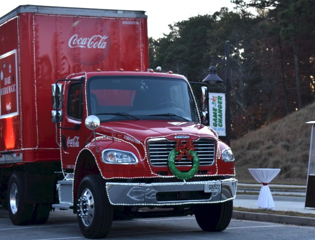 A decorated red Coca-Cola truck with a wreath on the front is parked outdoors, near a tall heater and a small table.