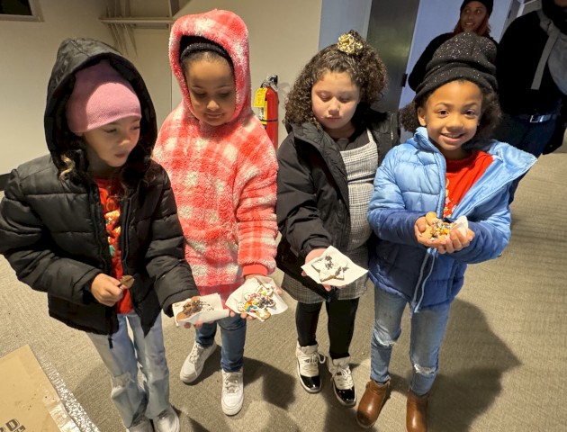 Four children in winter clothing are holding decorated treats in their hands, smiling indoors.