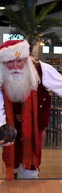 A person dressed as Santa Claus is bowling in a modern alley, with colorful seating and palm trees in the background.