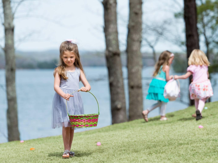 Young girls participating in an Easter egg hunt near a lake, each holding a basket, with trees in the background on a grassy hill.