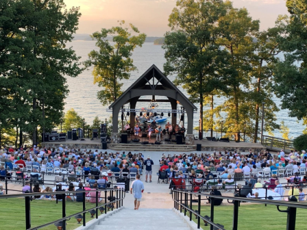 Outdoor concert with a crowd seated in front of a stage by a lake, surrounded by trees at sunset.