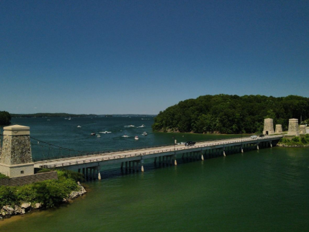 A suspension bridge spans over a body of water with boats, surrounded by lush green trees under a clear blue sky.