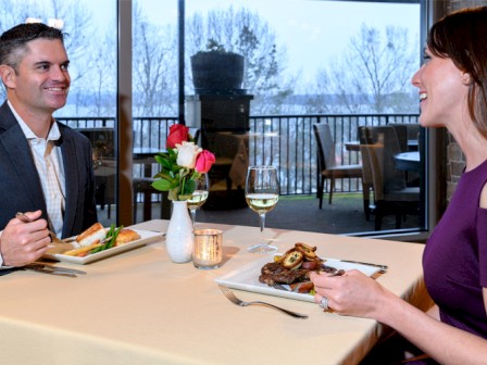 A couple is dining at a restaurant with wine, meals, and flowers on the table. A scenic view is visible through the large window.