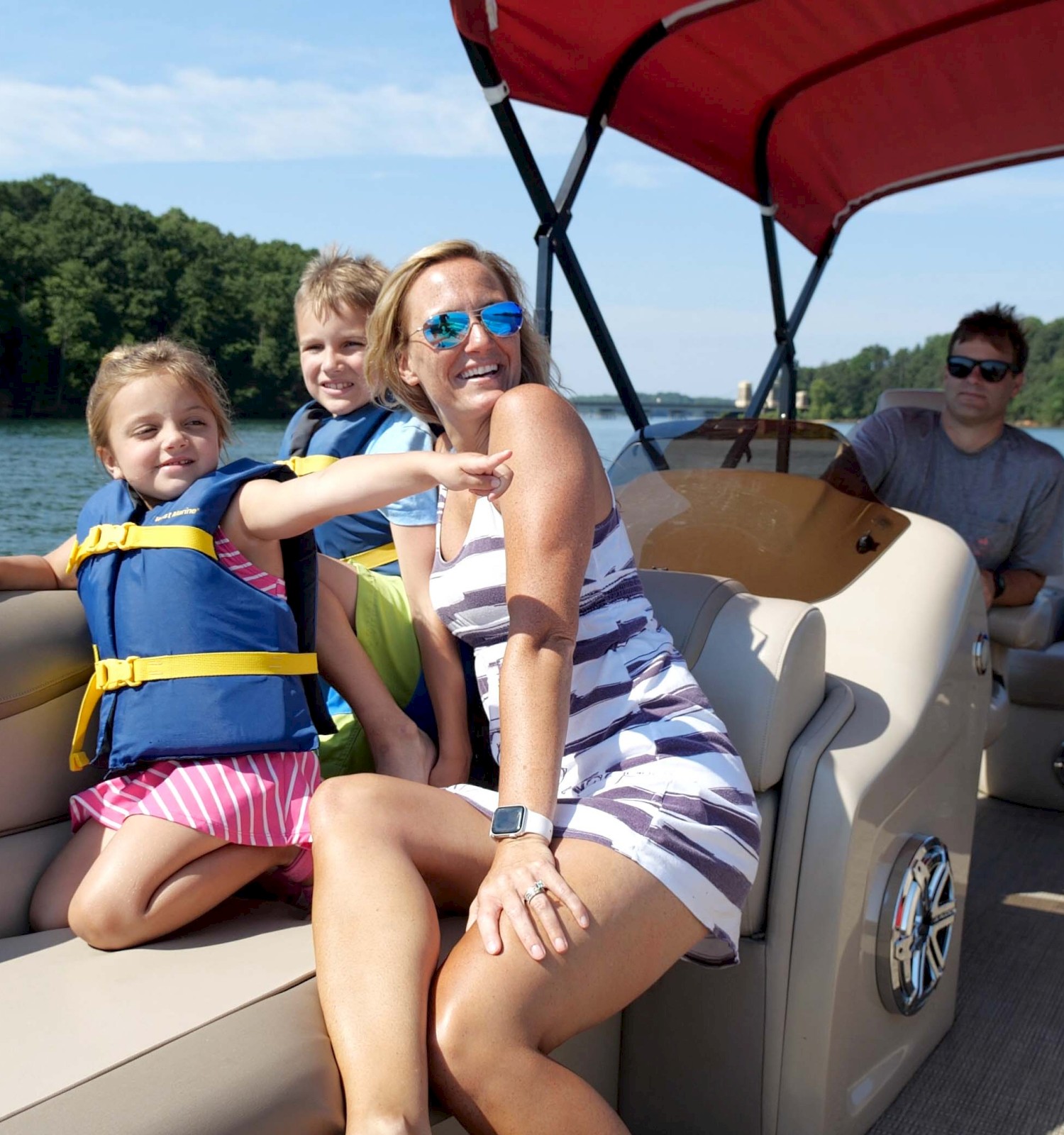 A group of people enjoying a sunny day on a boat, with children wearing life vests and an adult in sunglasses, amidst a scenic lake backdrop.