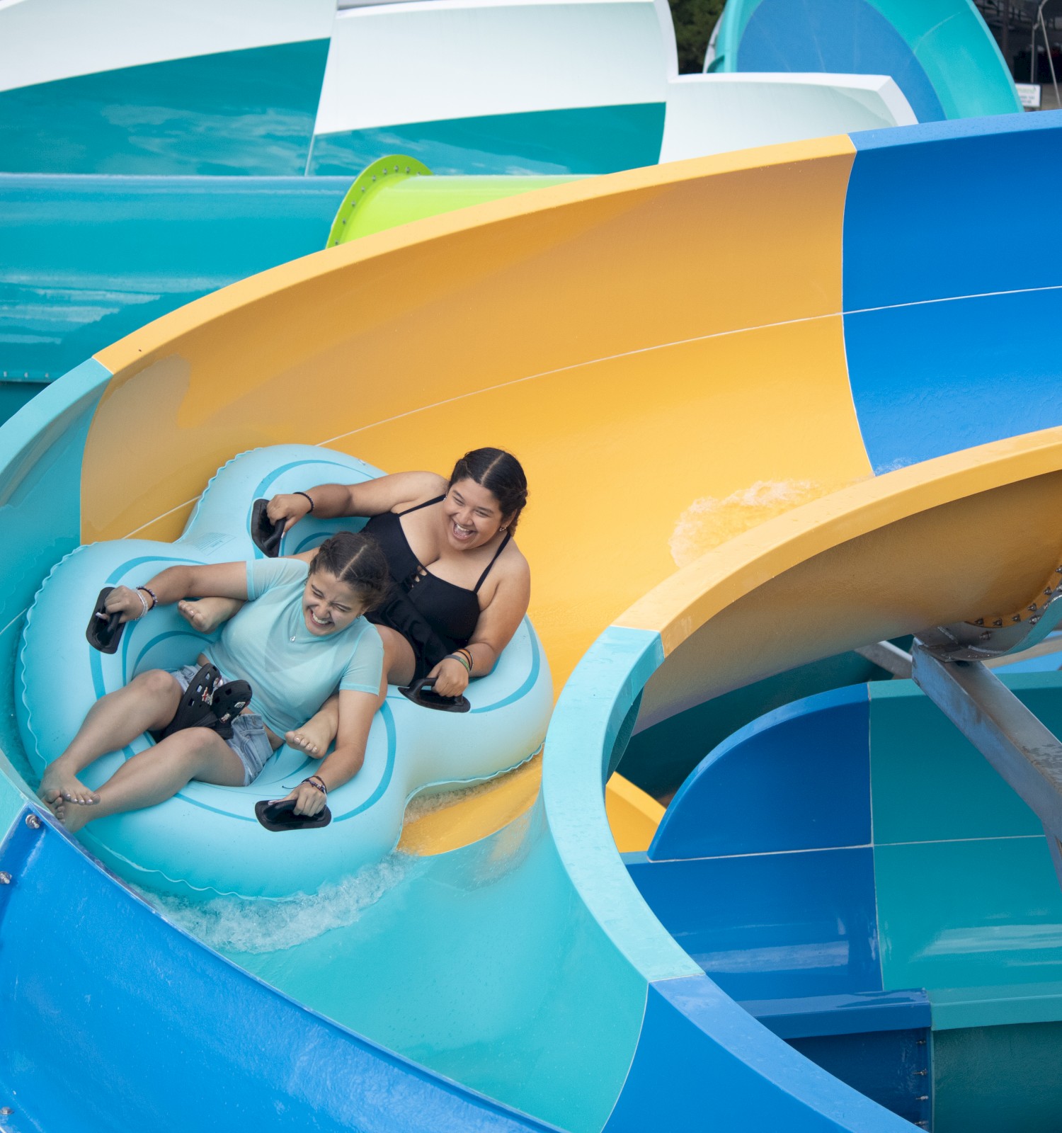 Two people are riding a blue inflatable raft down a colorful water slide at an amusement park.