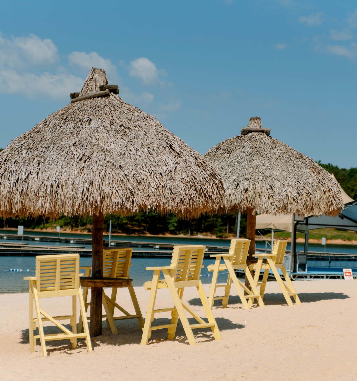 Sandy beach with thatched umbrellas and yellow chairs facing a calm body of water, surrounded by trees and a dock in the background.
