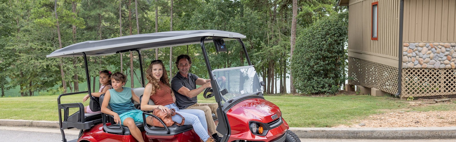 A family rides in a red golf cart near a house with trees in the background on a clear day.