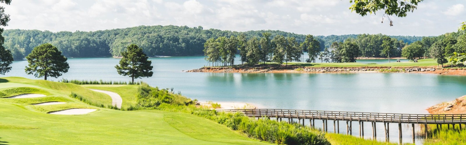 A scenic golf course with a bridge over water, surrounded by trees and lush greenery under a partly cloudy sky.