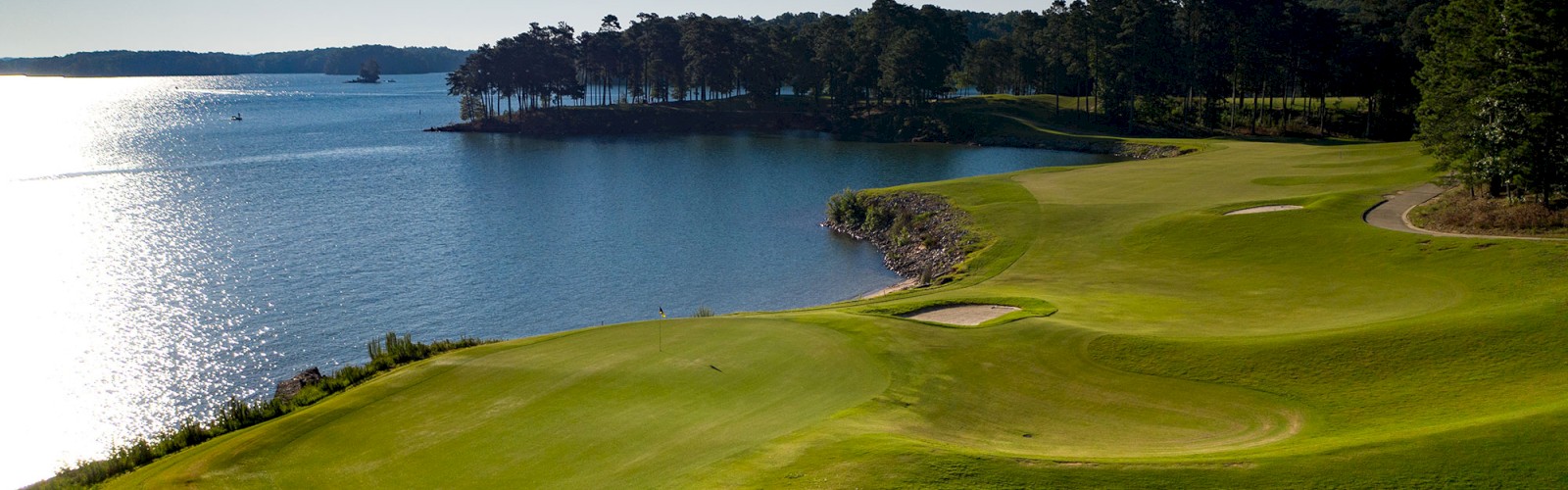 This image shows a serene golf course by a body of water with lush greenery, sand traps, trees, and a clear blue sky in the background.