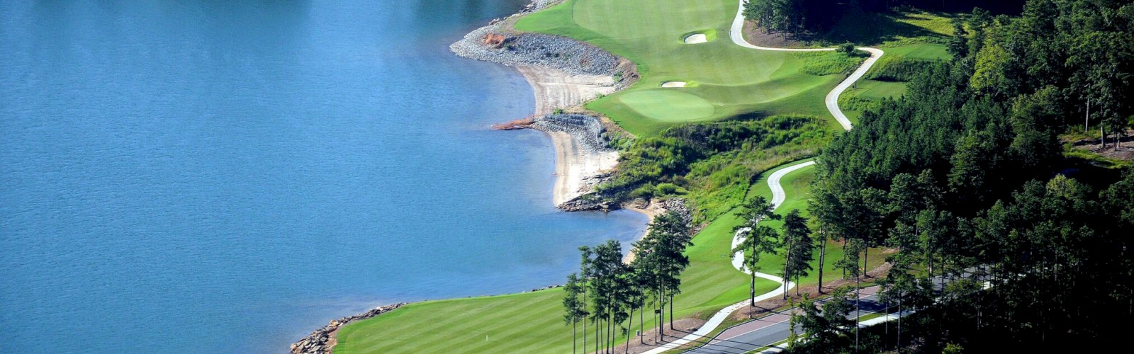 A scenic golf course is nestled by a lake, surrounded by lush greenery and trees, with winding paths and well-maintained fairways.