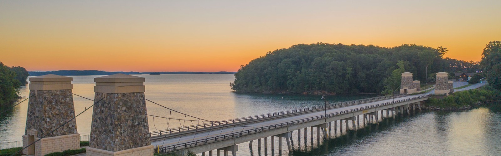 A stone-arched bridge spans a serene body of water at sunset, flanked by lush greenery and a clear sky.