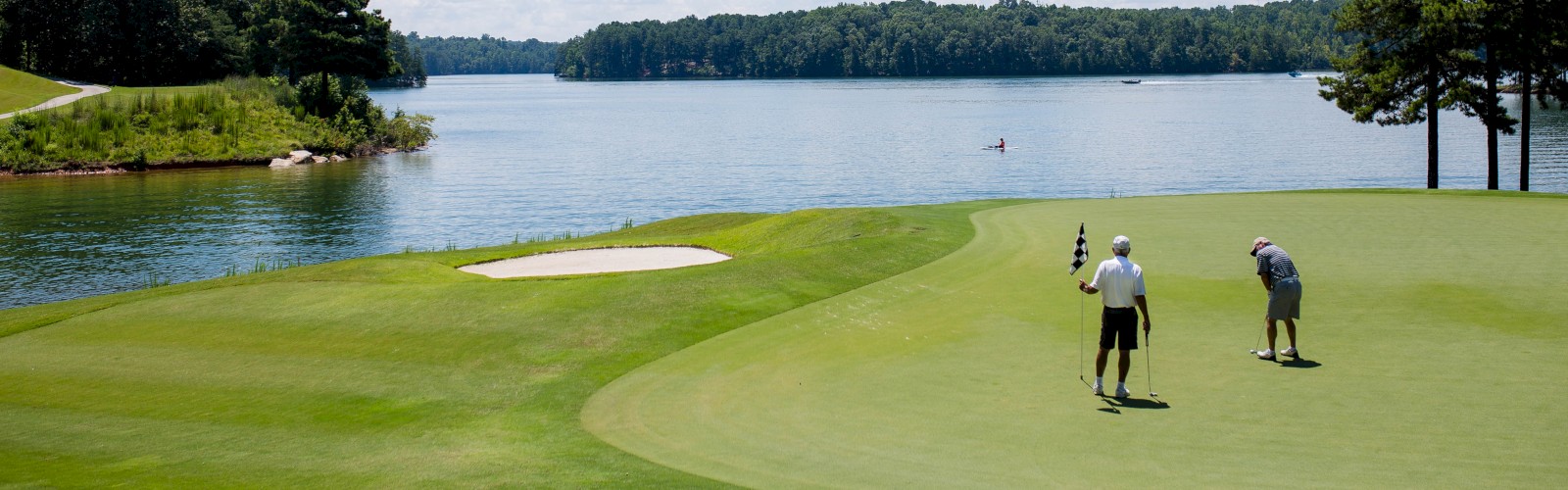 Two people are playing golf on a lush green course by a serene lake, under a partly cloudy sky.