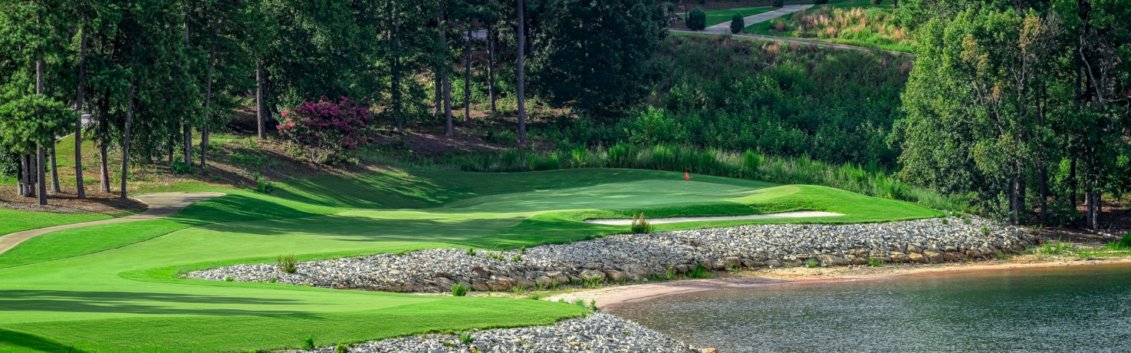 A scenic golf course with lush green fairways along a water body, surrounded by dense trees and a clear sky on a sunny day.