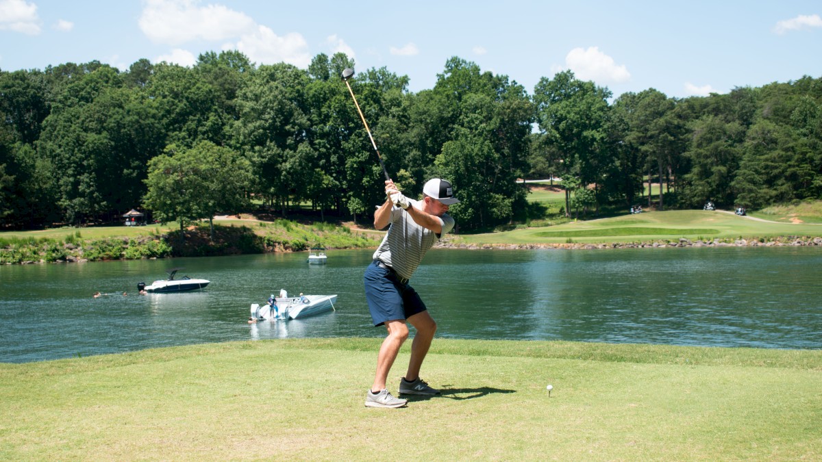 A golfer is swinging a club near a water hazard with trees in the background and a boat on the water.