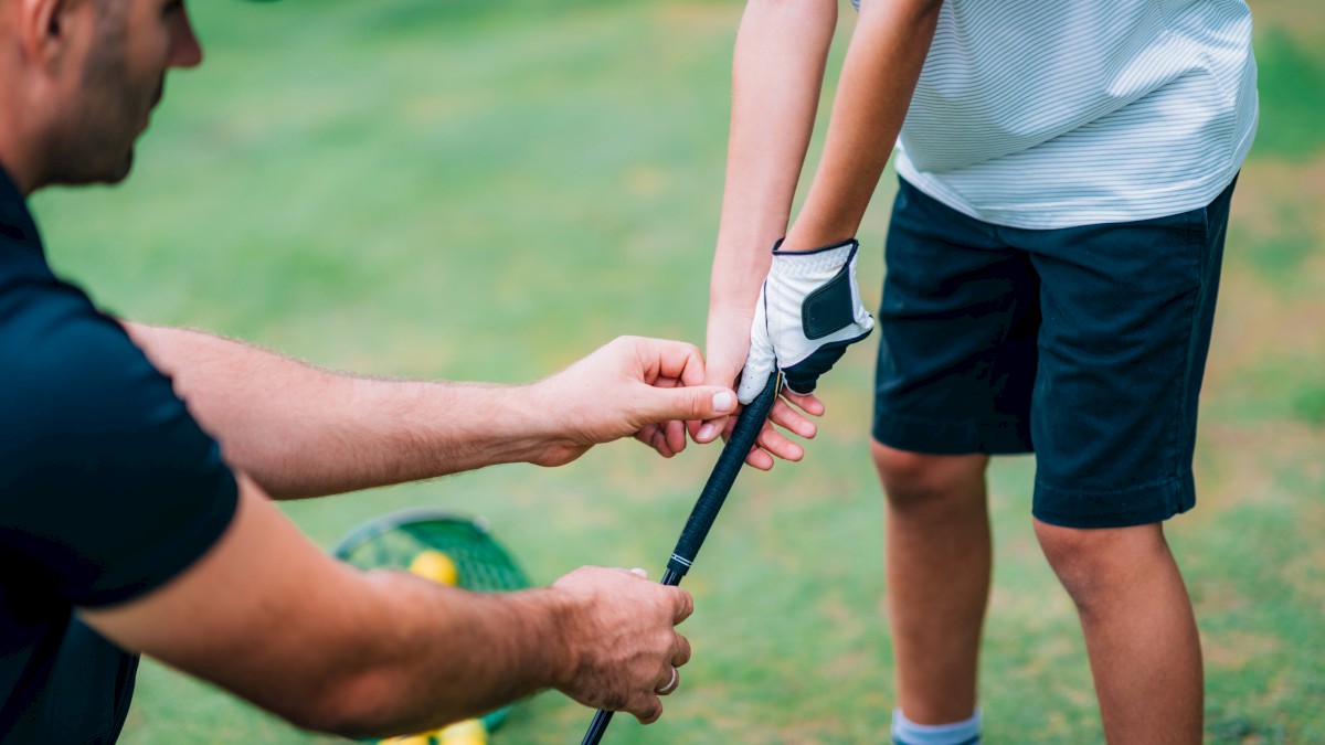 A person is teaching another how to hold a golf club on a green field, with a basket of golf balls nearby.