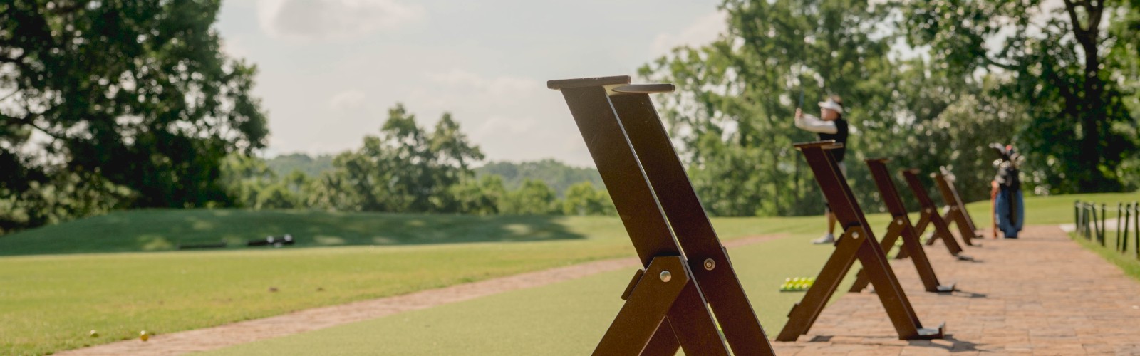 It's a golf driving range with practice tees, a basket of golf balls, and trees in the background under a clear sky.