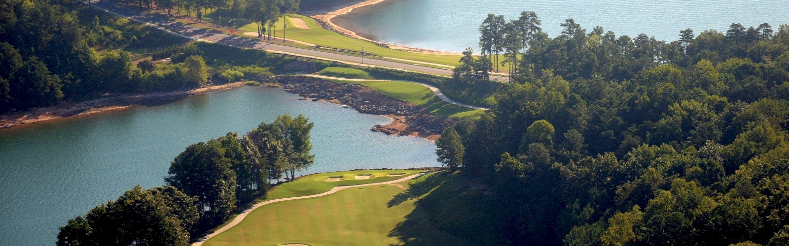 A scenic golf course overlooks a winding blue waterway, with lush green fairways, trees lining the banks, and a tranquil, sunlit landscape.