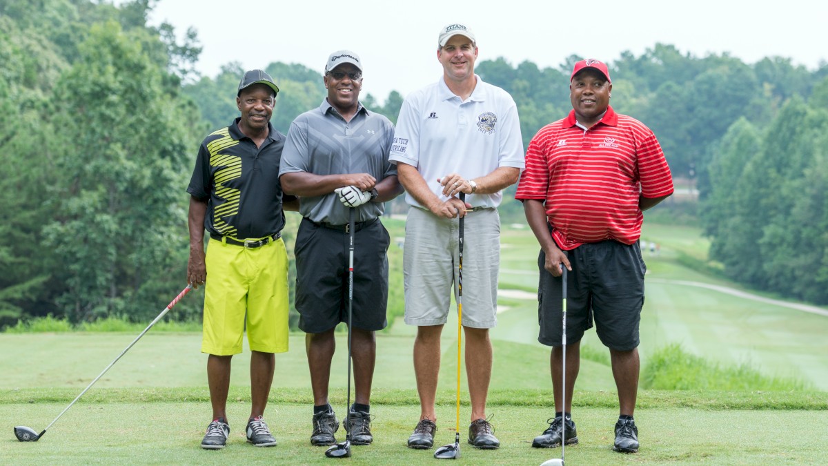 Four men are posing on a golf course holding clubs, with a grassy landscape and trees in the background.