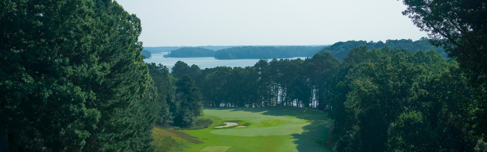 A golf course with a fairway surrounded by trees, and a body of water in the distance under a clear blue sky.
