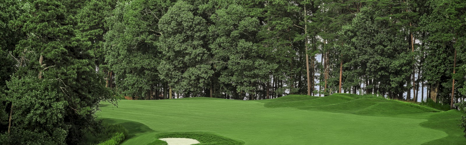 A scenic golf course with lush green fairways surrounded by trees and water in the background under a partly cloudy sky.
