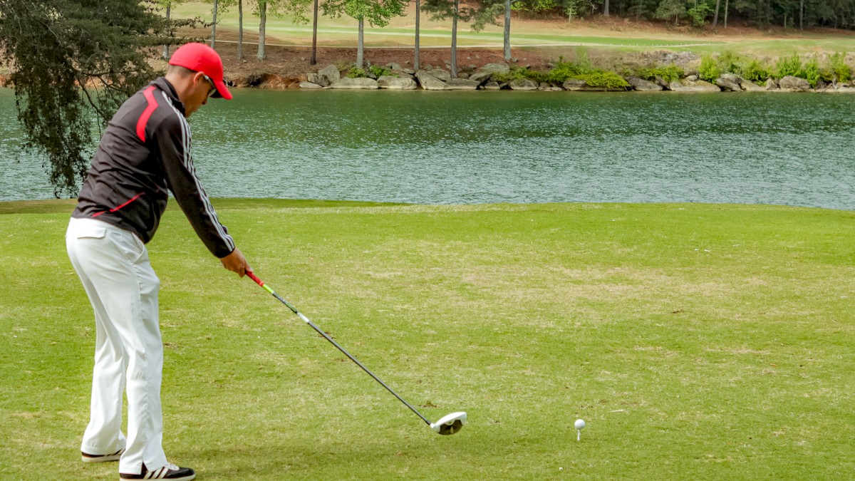 A person in golf attire is preparing to swing a club on a golf course near a body of water, surrounded by trees.